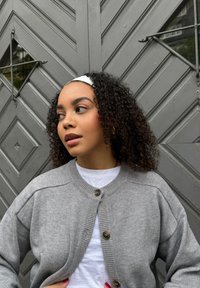 Cardigan gris à col rond, avec boutons et poignets côtelés, porté par-dessus une chemise blanche. L'arrière-plan présente une porte grise texturée.