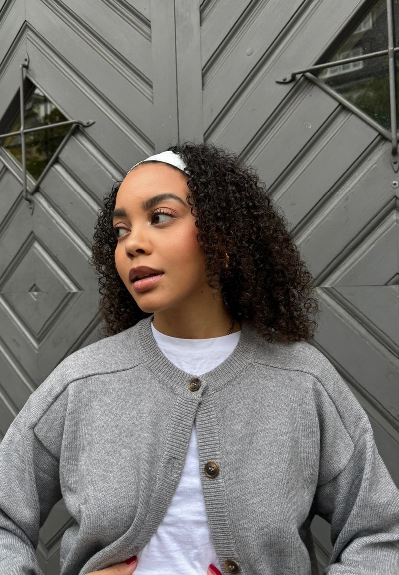 Cardigan gris à col rond, avec boutons et poignets côtelés, porté par-dessus une chemise blanche. L'arrière-plan présente une porte grise texturée.