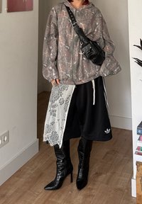 Veste matelassée grise à camouflage, sac bandoulière noir, écharpe en dentelle blanche, short noir et bottines à talons pointues noires, avec une finition texturée.