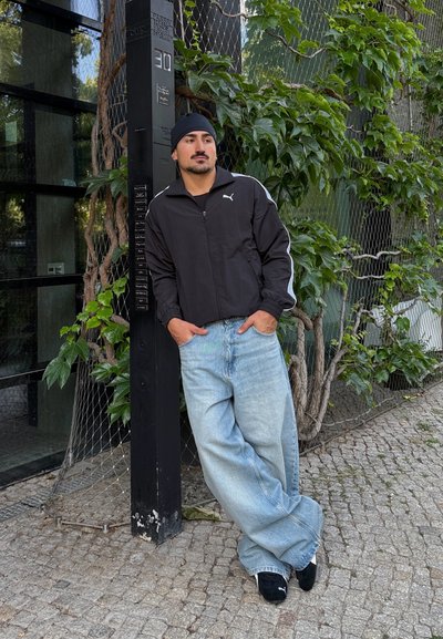 Chaqueta negra de Puma con cremallera y rayas blancas, pantalones vaqueros ligeros y holgados de color azul claro, y zapatillas negras, posando junto a una pared llena de hojas en un entorno urbano.