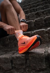Nike jooksujalats erksas oranžis, võrkpinnaga, musta tallaga ja punaste aksessuaaridega. Inimene reguleerib oranže lipsusid, samal ajal kui istub kivist treppidel.