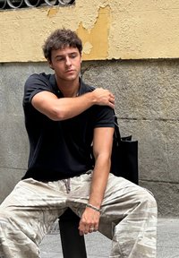 Fekete galléros póló, világos terepmintás nadrág, egy széken ül. Mellette fekete táska. Háttérben kőfal, kopott festékkel.