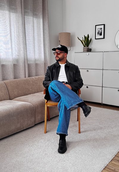 Hombre con gorra negra, gafas de sol, chaqueta de cuero, camisa blanca y vaqueros azules sentado con las piernas cruzadas en una silla de madera en un salón moderno.
