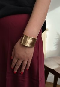 Un large bracelet manchette texturé en métal poli de couleur or, porté sur une main avec un vernis à ongles rouge, sur un fond en tissu bordeaux.
