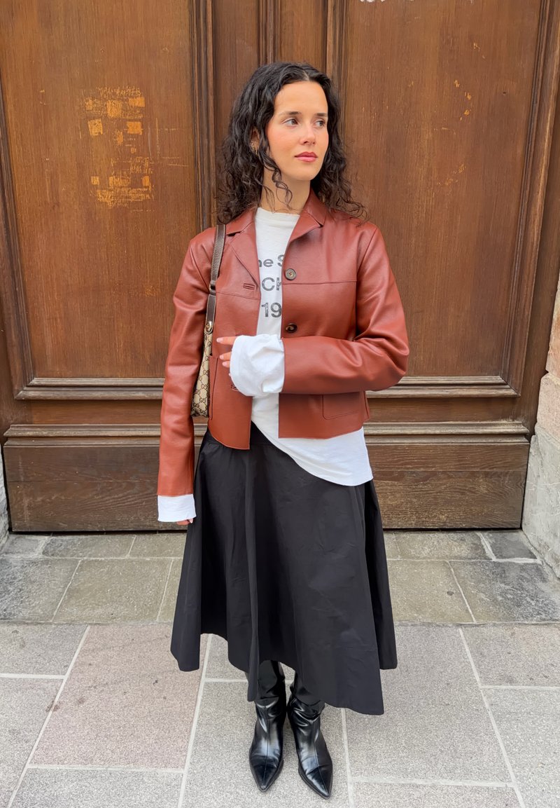 Femme aux cheveux bouclés portant une veste en cuir marron, une chemise blanche à manches longues, une jupe noire et des bottes noires, debout devant une porte en bois.
