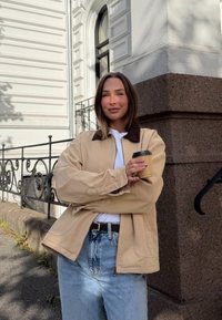 Giacca in tela beige con colletto marrone, indossata sopra una camicia bianca e jeans azzurri a vita alta, che tiene una tazza di caffè.