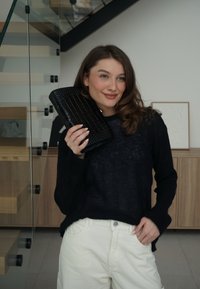 Νεαρή γυναίκα με καστανά μαλλιά κρατά μαύρο υφασμάτινο φάκελο κοντά στο πρόσωπό της, φορώντας μαύρο πουλόβερ και λευκό παντελόνι σε μοντέρνο εσωτερικό χώρο.