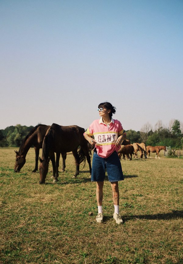 Maglietta polo rosa e gialla con logo "GANT", pantaloni corti in denim, sneakers bianche, in piedi in un campo erboso con cavalli marroni che pascolano.