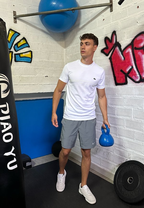 Jeune homme en chemise blanche et short gris tenant un kettlebell bleu dans une salle de sport avec un sac de frappe, un ballon d'exercice et des murs tagués.