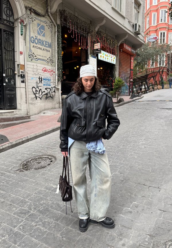 Jeune femme en veste en cuir noire et bonnet blanc en maille se tient sur une rue pavée près d'un café décoré de lumières de fête.