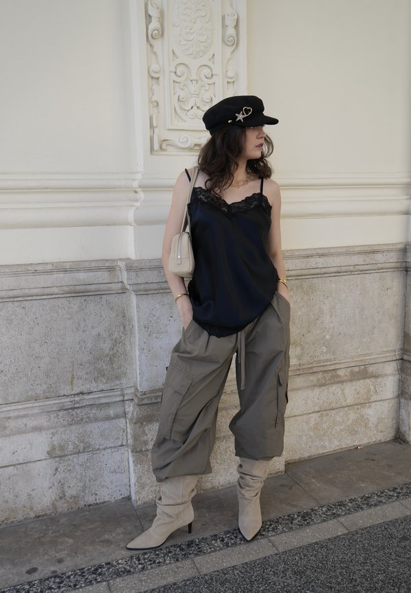 Femme portant un haut en dentelle noire, un chapeau noir, un sac beige, un pantalon cargo gris et des bottes beige amples, debout contre un mur de pierre.