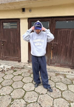 Sudadera gris con texto "PROHIBITED BERLIN", pantalones azules a rayas de corte holgado, gorra azul con logo blanco de "Yankees", zuecos negros, fondo de pavimento de piedra.