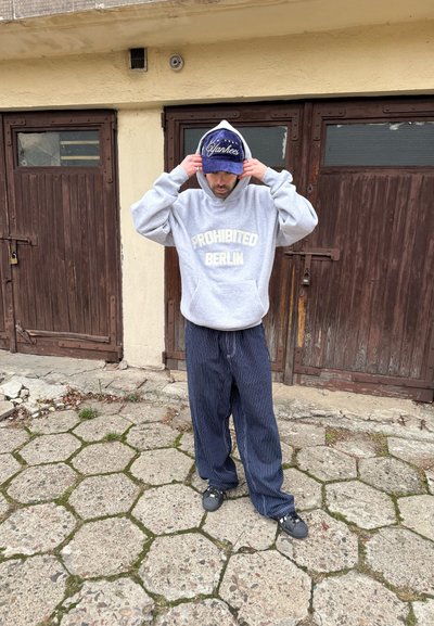 Sudadera gris con texto "PROHIBITED BERLIN", pantalones azules a rayas de corte holgado, gorra azul con logo blanco de "Yankees", zuecos negros, fondo de pavimento de piedra.