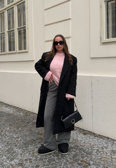 Abrigo negro de doble pecho, suéter de punto rosa, pantalones a rayas grises, bolso negro, zapatos negros. Texturas suaves, colores neutros, look casual.
