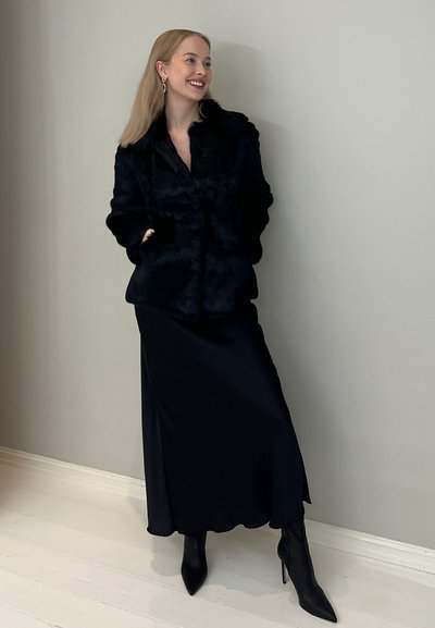 Chaqueta de piel sintética negra con un gran cuello, llevada sobre una larga falda de satén negra. Botas de tacón stiletto transparentes negras completan el look.