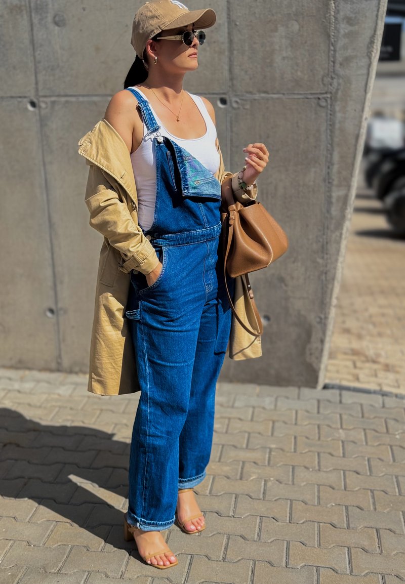 Salopette en denim d'un bleu profond, dotée de poches et de bretelles ajustables, superposée à un trench-coat beige. Complétée par un sac à main brun.