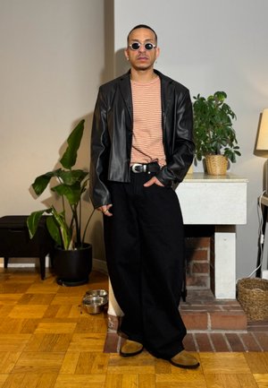 Hombre con gafas de sol redondas, chaqueta de cuero negra, camiseta de rayas, pantalones anchos negros y zapatos marrones de pie en el interior cerca de plantas y una chimenea.