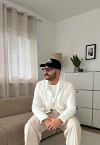 Fehér öltönyt és fekete sapkát viselő férfi ül egy bézs színű kanapén egy minimalista nappaliban, mögötte egy növény és egy állólámpa.