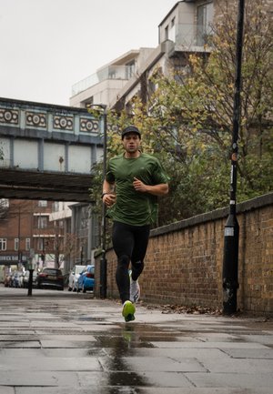 Hombre atlético corriendo sobre un pavimento mojado, vestido con una camiseta verde de manga corta, mallas negras y una gorra negra; complexión atlética fuerte.