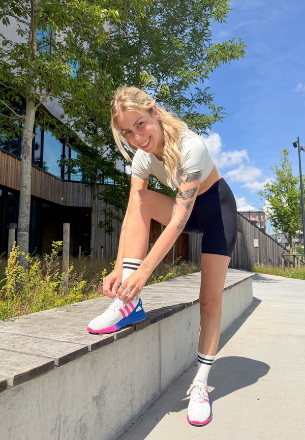 Joven mujer con ropa deportiva atándose zapatillas blancas y rosas al aire libre en un día soleado con árboles y un edificio moderno detrás.