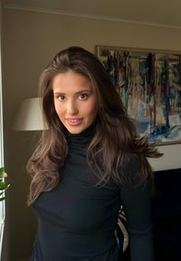 Młoda kobieta o długich brązowych włosach, ubrana w czarny golf, stoi wewnątrz przy oknie, za nią lampa i abstrakcyjny obraz.