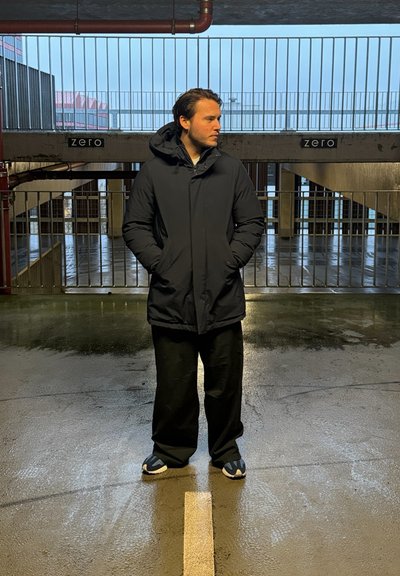 Parka negra impermeable con capucha, pantalones negros de corte holgado y sandalias azul oscuro; de pie sobre un suelo de hormigón mojado en un garaje.