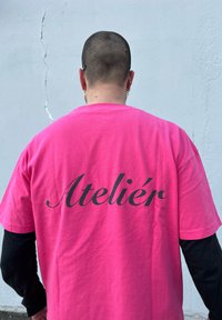 Ružová bavlnená tričko s veľkým čiernym kurzívou nápisom "Atelier" na chrbte. Krátke rukávy a voľný strih.