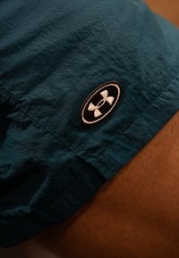 Teal lette shorts med en krøllet tekstur; har en sort oval lap med hvidt logo på kanten. Nærbillede.
