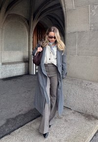 Femme portant un long manteau gris, un cardigan beige, un pantalon évasé gris, des lunettes de soleil et un sac à main marron foncé, marchant près d'arcs en pierre.
