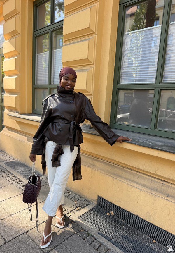 Jeune femme portant une veste en cuir marron, un pantalon blanc et un foulard, s'appuie sur la fenêtre d'un bâtiment jaune, tenant un sac à main matelassé.
