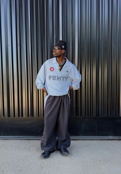 Joven con gorro negro, gafas de sol, chaqueta blanca de Fenty y pantalones oscuros holgados se encuentra de pie junto a una alta pared de metal negro. Con las manos en las caderas, mira hacia la izquierda.