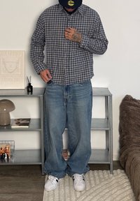 Chemise boutonnée à carreaux bleu et blanc, jean bleu ample et baskets blanches. Debout dans une pièce minimaliste avec des étagères et des objets décoratifs.