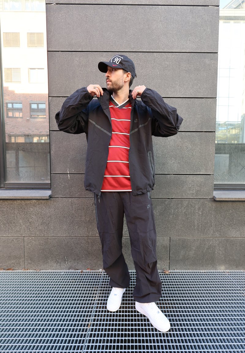 Homme portant une casquette noire, une veste noire et un pantalon, ajuste son col en se tenant sur une grille métallique devant un mur en pierre grise avec des fenêtres.