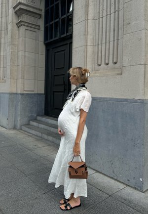 Vestido blanco texturizado con mangas cortas, un bolso marrón y sandalias negras. Cabello recogido en un moño, de pie contra una pared de piedra.