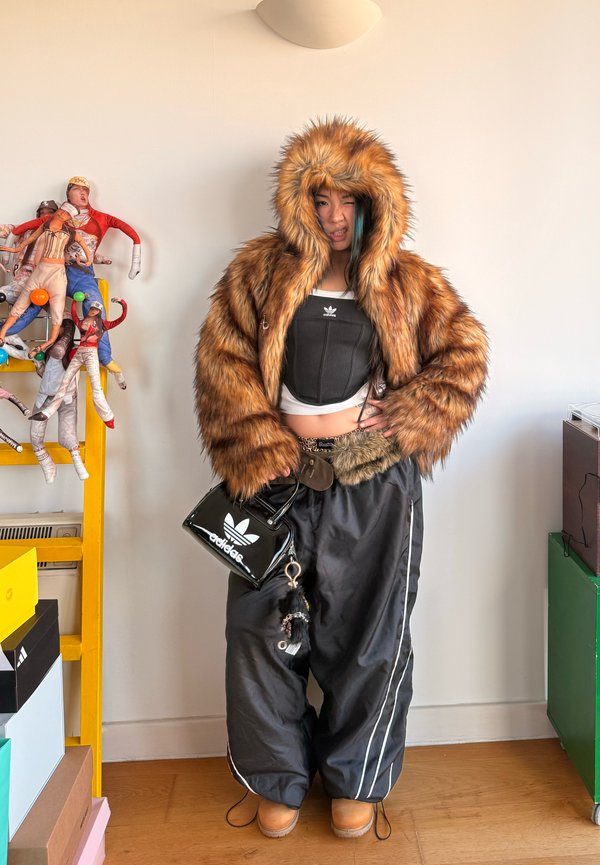 Asmuo, dėvintis didelę kailinę su gobtuvu striukę, juodą adidas trumpą viršutinį drabužį, plačias juodas kelnes ir smėlinės spalvos batus, laiko mažą juodą adidas krepšį patalpose.