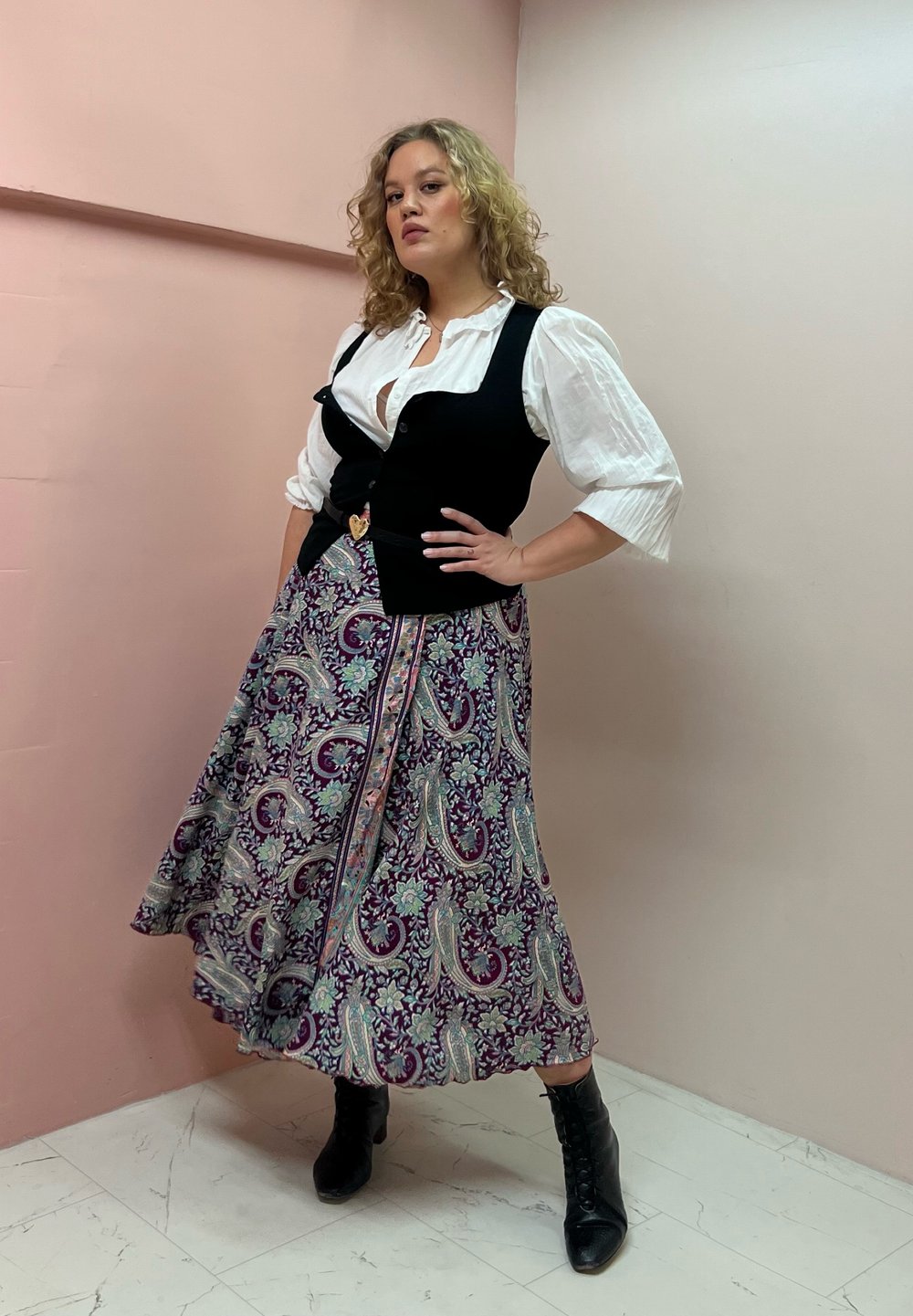 Schwarze Weste über einem weißen Hemd, kombiniert mit einem langen, gemusterten Rock in Lila, Türkis und floralen Designs. Schwarze Knöchelstiefel runden den Look ab.