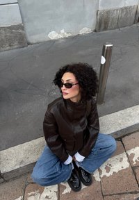 Brązowa skórzana kurtka, biała koszula z długim rękawem, niebieskie dżinsowe spodnie o szerokich nogawkach, czarne lakierowane buty i czarne okulary przeciwsłoneczne. Betoniasta tło.