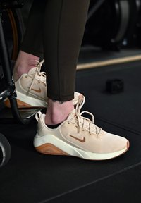 Chaussures de sport Nike beige avec une tige en mesh, des accents marron contrastants et une semelle en caoutchouc, placées à côté d'un fauteuil roulant et d'équipements d'entraînement.