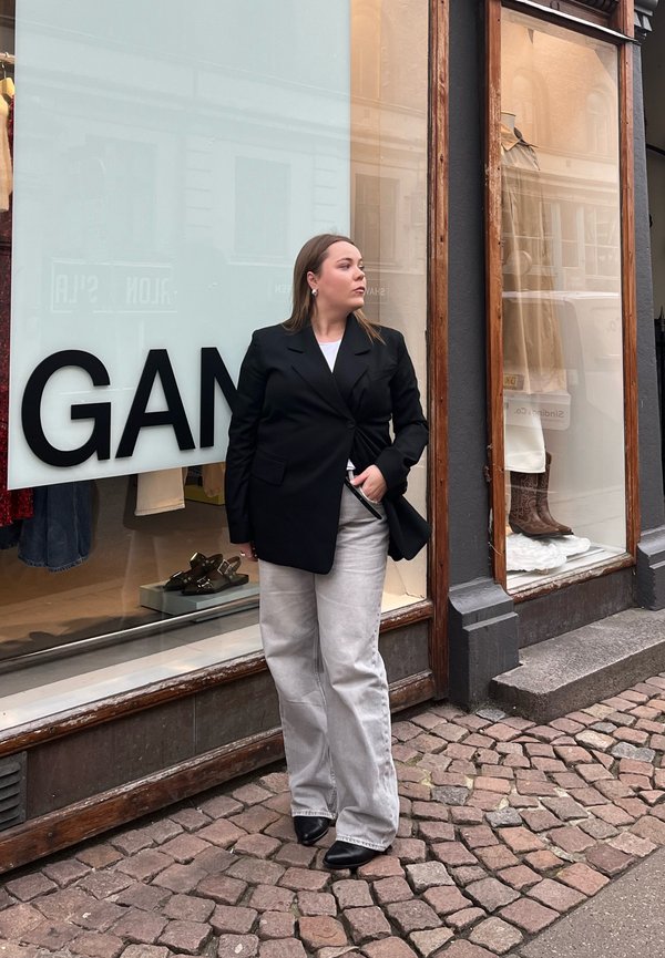 Črn blazer, bela majica in svetlo sivo široke hlače. Gladko blago, strukturirana zasnova in črne čevlje na blok peti. Ozadje iz kamnitih plošč.