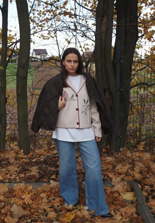 Jachetă subțire maro, cardigan bej cu broderie florală și blugi largi de culoare albastru deschis. Mediu natural cu frunze căzute.