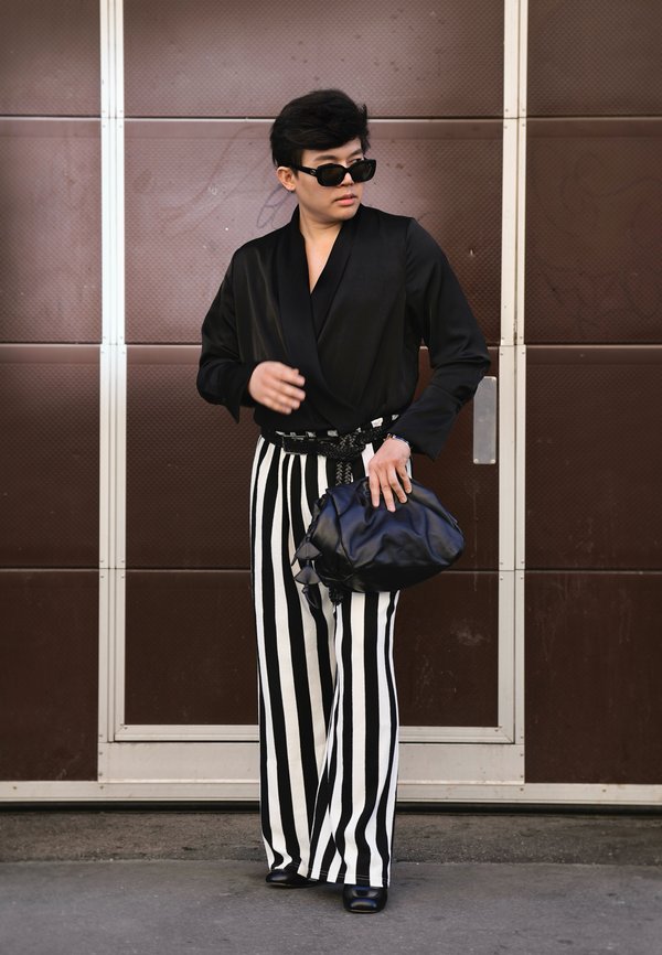 Personne portant un haut noir, un pantalon large à rayures verticales noires et blanches, des lunettes de soleil noires, tenant un sac à main noir, debout devant une porte marron.