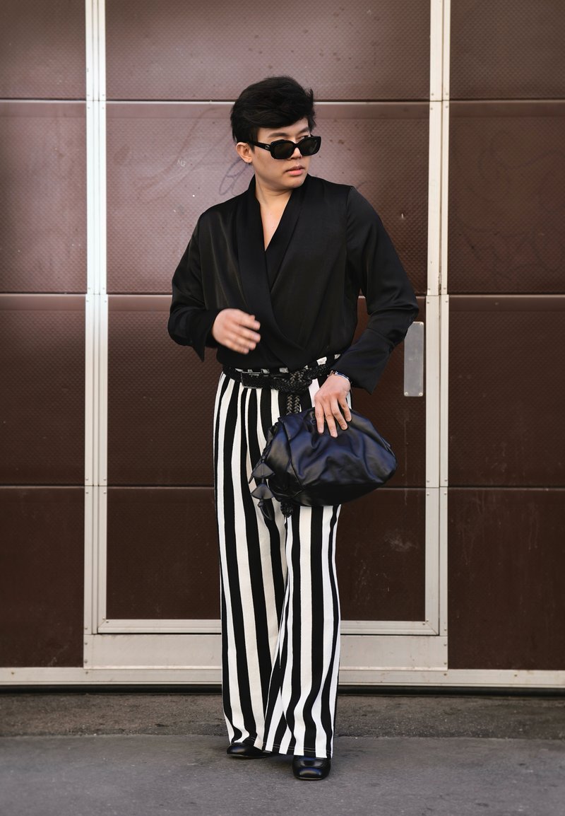 Personne portant un haut noir, un pantalon large rayé vertical noir et blanc, des lunettes de soleil noires, tenant un sac à main noir, debout devant une porte marron.