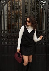 Rochie neagră fără mâneci peste o cămașă albă cu mânecă lungă, asortată cu o geantă texturată burgund și cizme negre până la genunchi. Ochelari de soare negri purtați.