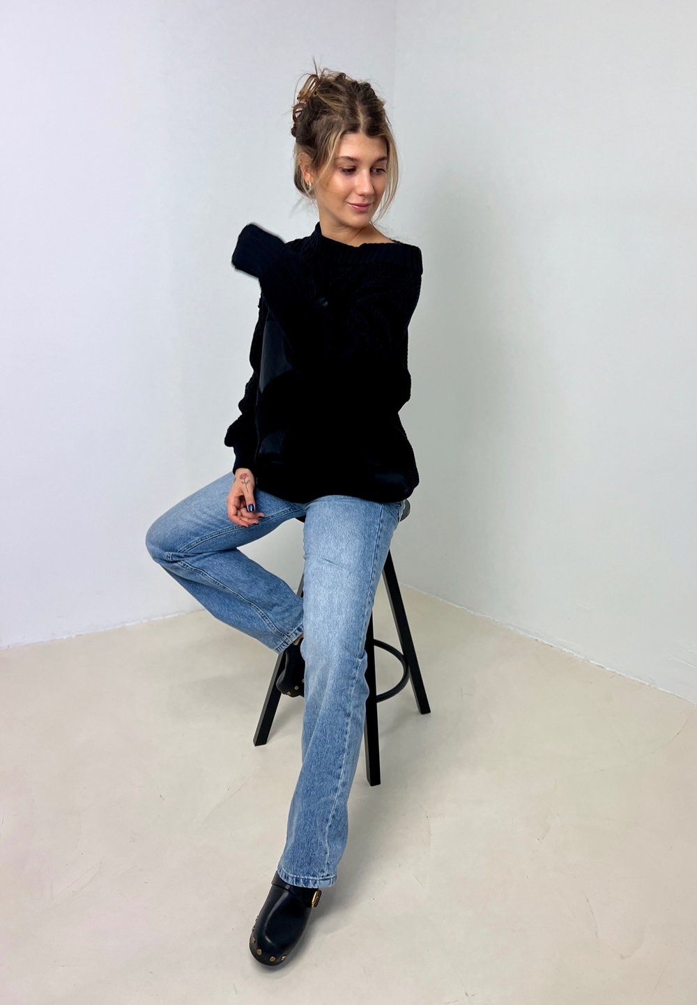 Suéter preto oversized com textura canelada, jeans azuis de perna reta e tamancos pretos sobre um banco contra um fundo branco liso.