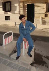 Jaqueta de ganga azul clara e calças de ganga à boca de sino combinadas, usadas com sapatos pretos de slip-on, sentado em uma barreira de construção num ambiente urbano.