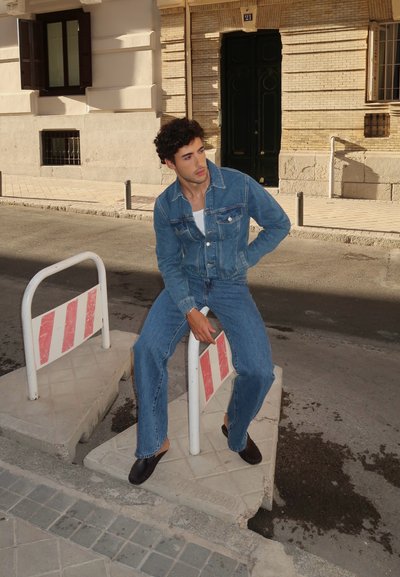 Chaqueta de mezclilla azul claro y jeans acampanados a juego, llevados con zapatos negros sin cordones, sentada sobre una barrera de construcción en un entorno urbano.