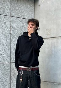 Svart hoodie med känguruficka, silverkedja och en metallspänne. Bärs med mörka jeans och nycklar hängande. Grå texturerad vägg i bakgrunden.