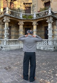 Homme portant une veste grise, un pantalon à carreaux et une casquette plate, debout dans une cour pavée, faisant face à un bâtiment historique orné de colonnes en pierre et de balustrades décoratives.