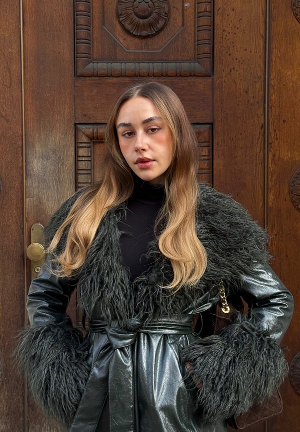 Nuori nainen, jolla on pitkä vaalea tukka, päällään musta nahkatakki, jossa on iso turkishuppu ja -hihansuut, seisoo veistetyn puuoven vieressä.