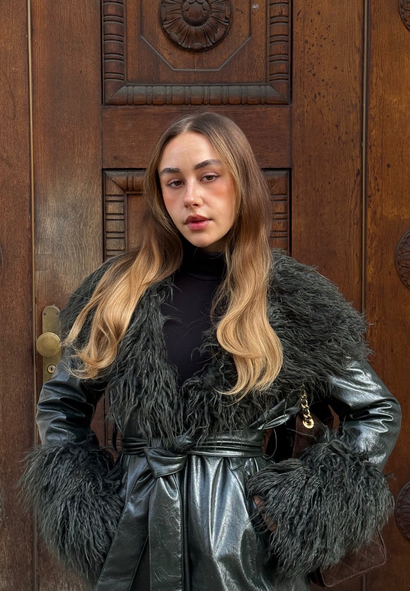 Jeune femme aux longs cheveux blonds portant un manteau en cuir noir avec un grand col et des poignets en fourrure, debout devant une porte en bois sculptée.